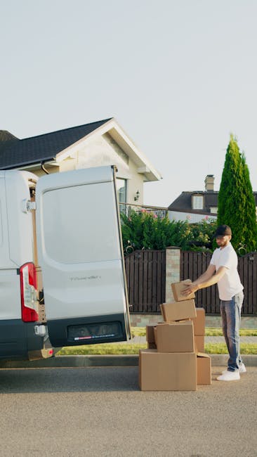 A man wearing a white t-shirt and jeans is standing beside an open white moving van parked on the street in a residential area, preparing boxes for a home relocation. He is handling cardboard boxes of